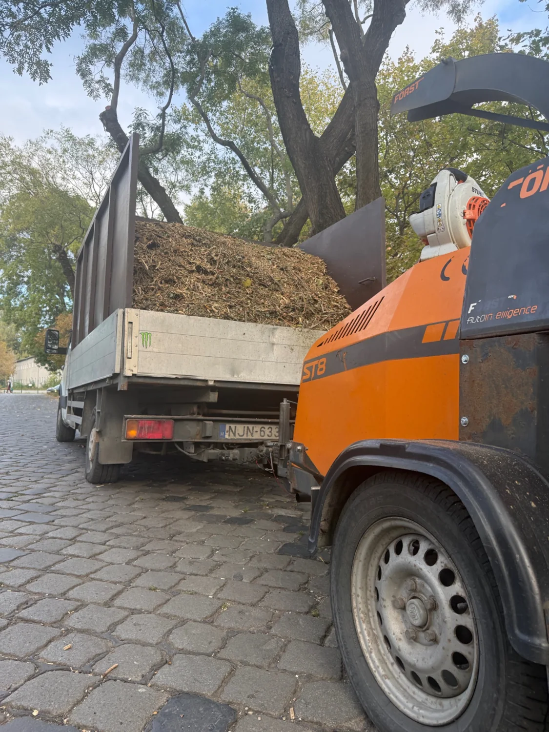 Narancssárga ágdaráló, mögötte aprítékkal megrakott jármű.